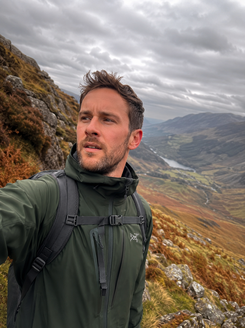 Man in a green waterproof hiking jacket and backpack on a mountain ridge above a valley — AtlasGears