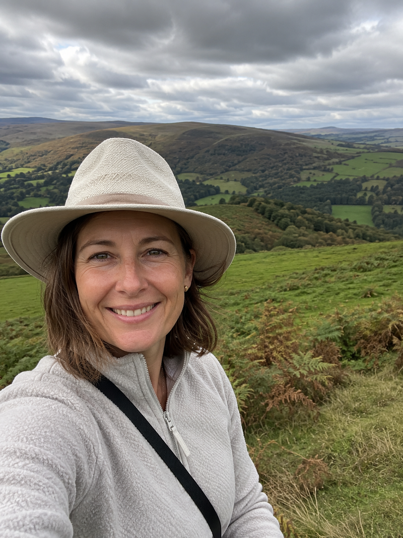 Smiling woman in a wide-brim sun hat and grey fleece on a green countryside hillside — AtlasGears
